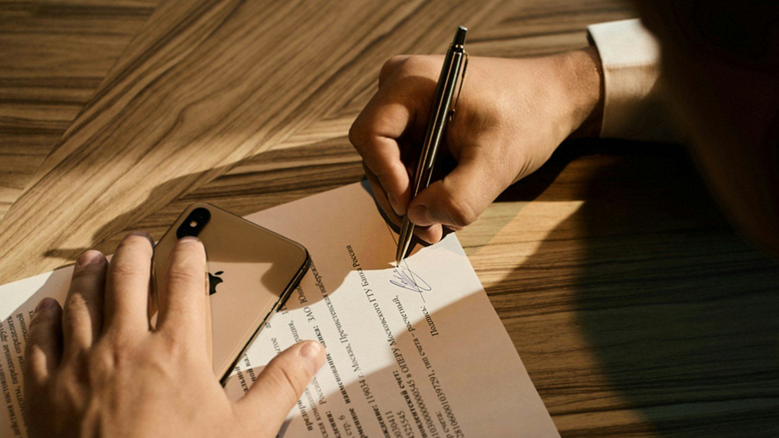 A hand signing a contract with a pen on a wooden desk, a phone resting on the page